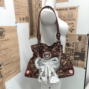 Peut inclure: Un sac à bandoulière marron orné d'un motif répétitif de couronnes dorées, agrémenté d'un nœud en dentelle blanche, de chaînes argentées et d'un emblème circulaire central avec des strass. Il possède des anses et des côtés en similicuir marron.
