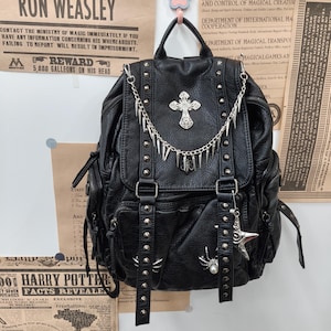 Può includere: Uno zaino nero testurizzato con borchie metalliche argentate, un emblema a croce argentato decorativo e una catena pendente con ciondoli a punta. La borsa include due cinghie verticali con fibbia sulla patta anteriore e ciondoli a ragno.