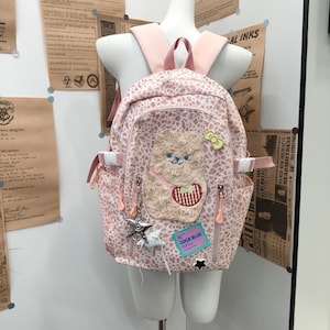 May include: A light pink backpack with a white leopard print pattern. The bag features a large, textured, beige plush bear appliqué, a small green bow pin, a silver star charm, and a rectangular blue tag with the text COCA BLUE printed on it.