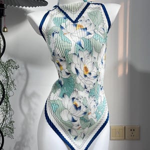 Könnte beinhalten: Ein ärmelloses, plissiertes Neckholder-Top mit weißer Basis und einem großflächigen Blumenmuster in Grün-, Blau- und Gelbtönen. Das Kleidungsstück hat einen hohen Ausschnitt und einen spitz zulaufenden Saum, beide mit dunkelblauem Rand.