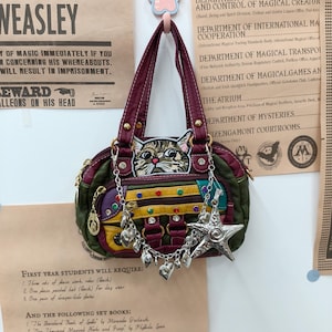 Könnte beinhalten: Eine mehrfarbige Handtasche mit einem Katzen-Aufnäher, goldfarbenen Beschlägen und einem silberfarbenen Sternanhänger. Die Tasche besteht aus bordeauxroten, grünen, gelben und violetten Einsätzen, die mit bunten Strasssteinen verziert sind.