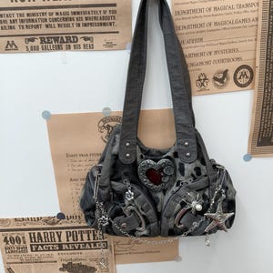 Può includere: Una borsa a spalla grigio scuro con stampa leopardata, caratterizzata da un prominente emblema argentato a forma di cuore con centro rosso, molteplici catene metalliche argentate, spille da balia, un ciondolo a ragno e uno a stella.