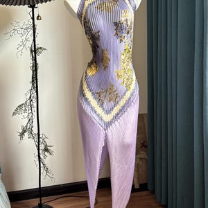 May include: A sleeveless, pleated midi dress in a light lavender hue, featuring a high neckline and a scarf-like floral print in shades of yellow, blue, and brown. The garment has a pointed, asymmetrical hemline that creates a distinct silhouette.