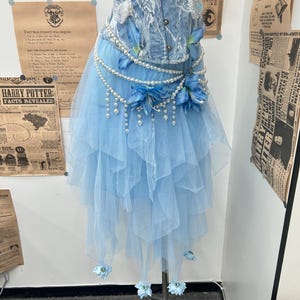 May include: A light blue denim corset top with white lace overlays paired with a layered tulle skirt. The ensemble is accented with strings of white pearls, blue fabric flowers, and small blue floral embellishments at the hem.