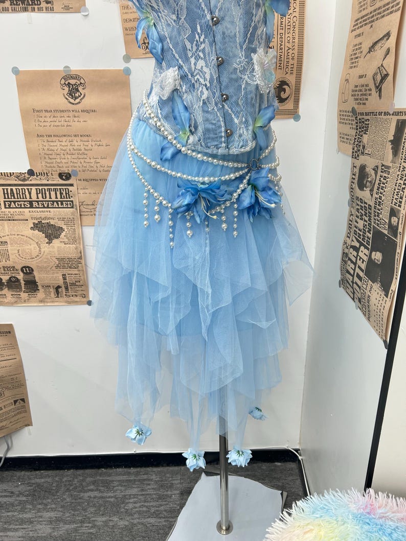 May include: A light blue denim corset top with white lace overlays paired with a layered tulle skirt. The ensemble is accented with strings of white pearls, blue fabric flowers, and small blue floral embellishments at the hem.