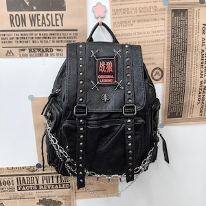Peut inclure: Un sac à dos noir à la texture grainée, orné de clous métalliques argentés sur les sangles et le rabat. Un écusson rectangulaire portant le texte rouge et les caractères chinois 战狼, ainsi que les mots ORIGINAL LEGEND, est fixé par des épingles.