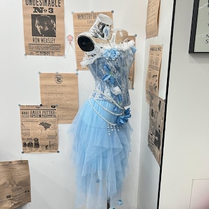 May include: A light blue corset top and layered tulle skirt set on a mannequin. The corset features white lace overlays, blue fabric flowers, and a draped pearl chain accent. The skirt has an asymmetrical hemline with small blue flower accents.