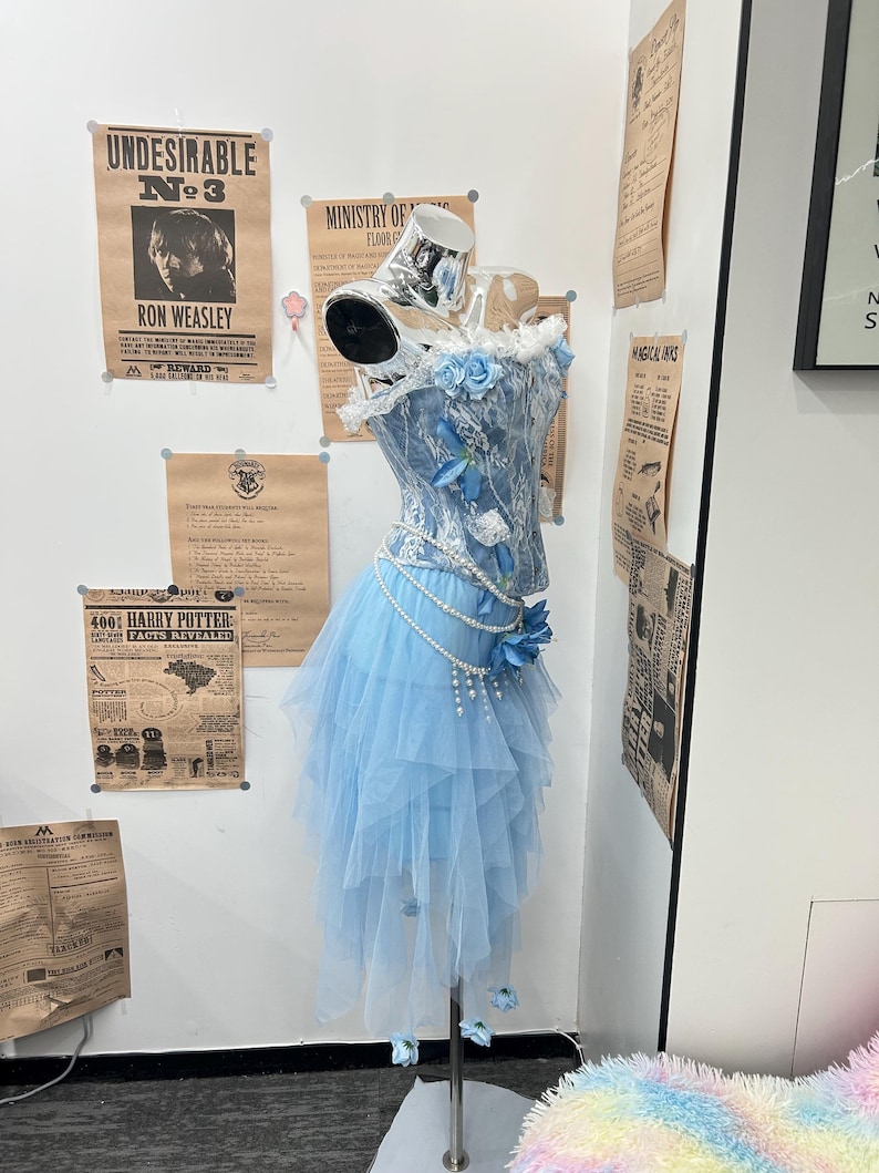 May include: A light blue corset top and layered tulle skirt set on a mannequin. The corset features white lace overlays, blue fabric flowers, and a draped pearl chain accent. The skirt has an asymmetrical hemline with small blue flower accents.