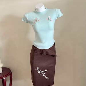 May include: A light mint green short-sleeved ribbed knit top featuring two embroidered butterflies on the chest, paired with a dark brown knee-length skirt that has a matching fabric belt and a white floral branch embroidery on the lower front.