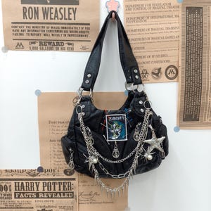 Pode incluir: Uma bolsa de ombro preta com várias correntes de metal prateado, pingentes pendentes de estrela e aranha, e um patch retangular com texto em japonês e um gráfico de caveira. A bolsa inclui tachas prateadas, zíperes e alfinetes.