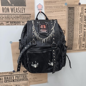 May include: A black faux leather backpack featuring silver-toned metal studs, a chain draped across the top, and safety pins. A rectangular patch with red and white text reads "战狼 ORIGINAL LEGEND". Two silver spider ornaments with white pearls are attached.