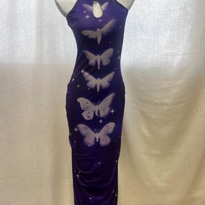 May include: A sleeveless, floor-length purple dress featuring a repeating vertical pattern of white moths and small four-pointed stars. The garment has a high halter neckline with a central teardrop-shaped cutout and a fitted, ruched silhouette.