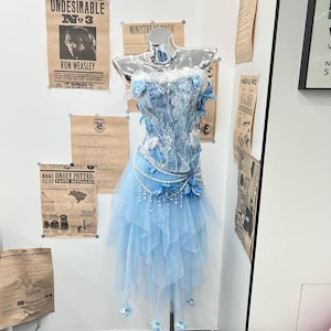 May include: A light blue corset top and matching layered tulle skirt set on a mannequin. The corset features white lace overlays, pearl strands, and blue fabric flowers. The skirt has a handkerchief hemline adorned with small blue flowers.
