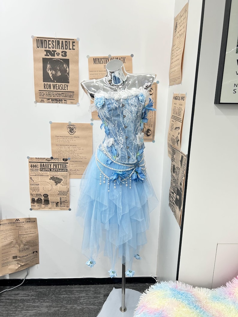 May include: A light blue corset top and matching layered tulle skirt set on a mannequin. The corset features white lace overlays, pearl strands, and blue fabric flowers. The skirt has a handkerchief hemline adorned with small blue flowers.