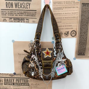 Könnte beinhalten: Eine braune Kunstleder-Schultertasche mit Leopardenmuster-Einsätzen, einem zentralen Schnallenriemen und einem goldenen Sternaufnäher mit roter Mitte. Die Tasche ist mit silbernen Ketten, einer Spinnenbrosche mit Perle, einem Sternanhänger und einem blauen Etikett mit der Aufschrift COCA BLUE verziert.