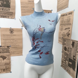 Puede incluir: Una blusa sin mangas de color azul claro con cuello alto, textura de punto y un estampado gráfico de un pájaro colorido posado en una rama con hojas rojas. La prenda se exhibe sobre un maniquí blanco.