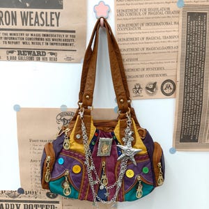 Peut inclure: Un sac à main multicolore composé de panneaux marron, violets, jaunes et bleu sarcelle, orné de boutons jaunes et bleus, d'une breloque en forme d'étoile argentée et de chaînes métalliques pendantes agrémentées de petites étoiles sur le devant.