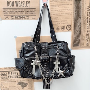 Puede incluir: Un bolso de mano negro con una correa central vertical con tachuelas y hebilla, dos bolsillos laterales y herrajes plateados. El bolso está adornado con cadenas plateadas colgantes, un dije en forma de estrella y una cruz con cristales.