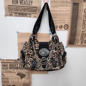 May include: A leopard print shoulder bag featuring black leather-like trim, a central silver buckle plate with the text GOOD GIRL, dangling silver chains, and two black star-shaped patches with white stitched outlines on the front flap.