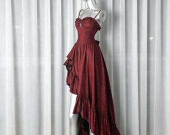 May include: A dark red high-low dress with a shimmering finish, featuring thin spaghetti straps, a ruffled neckline, and a silver cross pendant at the center. The skirt has layered ruffles and a black underskirt, creating a dramatic silhouette.