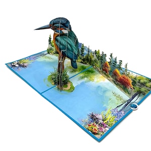May include: A 3D pop-up card depicting a kingfisher bird above a lake scene. The card has a blue border and features a detailed watercolor landscape with trees and colorful flowers. The card is a unique design.