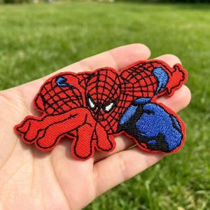 May include: An embroidered fabric patch featuring a red and blue superhero character with a black web pattern. The character is depicted in a crouching pose with white eyes, black outlines, and a textured surface created by dense thread stitching.
