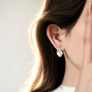 May include: A silver-toned earring with a feather design, featuring small clear stones and a single pearl. The earring is worn on the ear of a person with long, dark brown hair.
