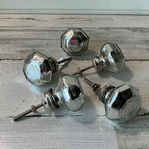 May include: Five faceted glass cabinet knobs with silver hardware. The knobs have a reflective, mercury glass appearance and are arranged on a light-colored, weathered wooden surface. The knobs are likely for furniture restoration or home decor projects.