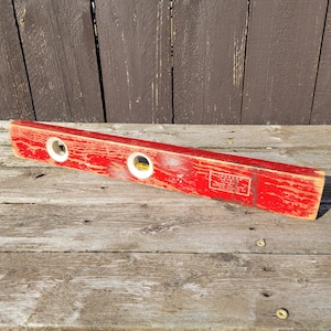 May include: A vintage red wooden level with two circular viewing windows. The level has a faded paint finish and a rectangular label with text. The level is approximately 61 cm long.