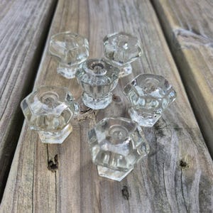 May include: A set of six clear glass cabinet knobs, each featuring a multi-faceted, hexagonal shape with a central circular indentation. The knobs are arranged in a cluster, showcasing their transparent, reflective surfaces and geometric design.