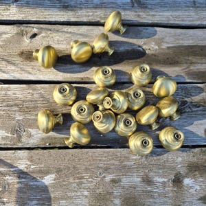 May include: A collection of brass mushroom-shaped cabinet knobs scattered on a weathered wooden surface. The knobs have a rounded top and a cylindrical base, with a screw hole visible on the front. The knobs are a warm gold color.