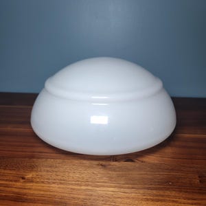 May include: A white, opaque glass lampshade with a rounded, bowl-like shape. The shade has a smooth, glossy surface and a slightly raised, circular band near the top. It sits on a dark wooden surface.