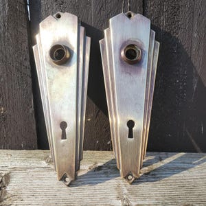 May include: Two vintage silver-toned door handle backplates. Each plate has a keyhole, a circular opening, and a decorative Art Deco fan design. The plates are mounted on a weathered wooden surface.