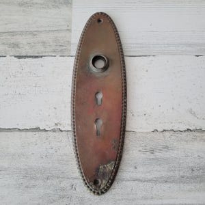 May include: An antique, oval-shaped door plate with a tarnished bronze finish. The plate features a circular opening at the top, two keyhole slots, and a decorative beaded border. The metal shows signs of age and wear.