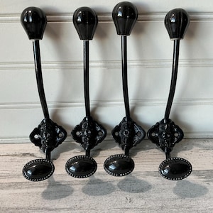 May include: Four black metal coat hooks with a decorative design. Each hook has a curved stem, a decorative base, and a rounded top. The hooks are mounted on a white and gray wooden surface.