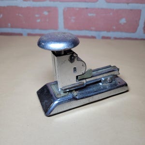 May include: Vintage silver stapler with a rounded top and a rectangular base. The stapler has a lever and a metal construction, showing signs of wear. The background features a brick wall.
