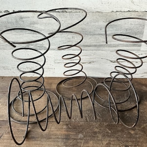 May include: Three antique metal spring sculptures with a dark, aged finish. Each sculpture features a circular top supported by a spiral spring base. The bases are attached to a circular metal plate. The sculptures are displayed on a wooden surface.
