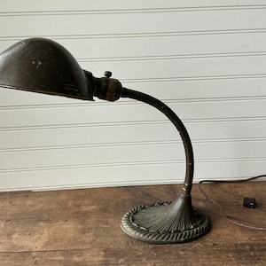 May include: An antique desk lamp with a dark, domed shade and a flexible, gooseneck arm. The base has a decorative, circular design. The lamp is made of metal and has a vintage aesthetic. The power cord is visible.