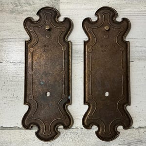 May include: Two antique bronze-colored metal door plates with ornate, symmetrical designs. Each plate has a decorative top, a rectangular center, and a stylized bottom. The plates have a vintage aesthetic, with visible wear and tear.