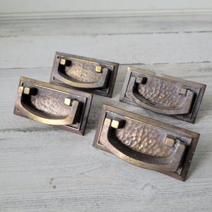 May include: Four antique bronze drawer pulls with a hammered texture and a curved handle. The pulls have a rectangular backplate and are set against a light wood background. These vintage hardware pieces are ideal for furniture restoration.