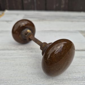 May include: A vintage door knob set featuring two round, dark brown, speckled ceramic knobs connected by a central metal spindle. The knobs have a glossy, reflective finish with visible signs of age and wear on the metal components.