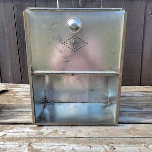 May include: Vintage metal wall-mounted cabinet with a silver finish. The cabinet features a diamond-shaped ventilation pattern and a round knob. The cabinet has a shelf and is likely for storage. The cabinet is made of metal and has a rectangular shape.