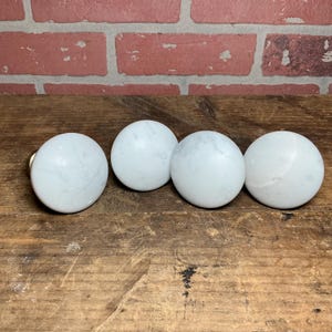 May include: Four white marble cabinet knobs with subtle gray veining are arranged on a wooden surface. One knob has a gold-colored base. The background features a red brick wall. These knobs are suitable for furniture restoration or home decor projects.