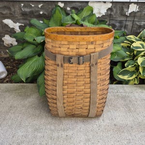 May include: A tall, oval-shaped woven wood basket featuring a light brown finish. The basket is secured with a wide, tan-colored fabric strap that wraps around the exterior and fastens with a metal buckle, providing a structured appearance.