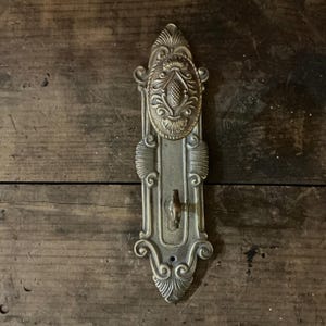 May include: An antique brass door handle with detailed floral and scrollwork. The handle has a keyhole and is mounted on a wooden surface. The design is symmetrical and classic, with a vintage aesthetic.
