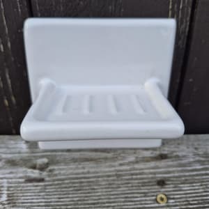 May include: A white ceramic soap dish featuring a rectangular wall-mounting plate and a protruding shelf with four parallel drainage grooves. The surface is smooth and glossy, designed to hold a bar of soap securely while allowing water to drain.