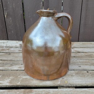May include: A round, brown ceramic jug with a glossy, reflective glaze. The jug features a wide, bulbous body, a short, narrow neck with a small spout, and a single curved handle attached to the side. The surface shows subtle color variations.