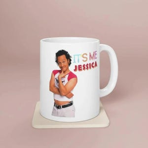May include: White ceramic mug featuring a colorful image of a man with the text "IT'S ME JESSICA". The mug has a white handle and is sitting on a square, off-white coaster.