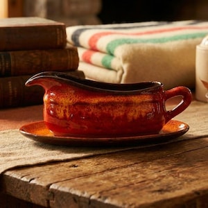 May include: A red and brown ceramic gravy boat with a matching saucer on a wooden table. The gravy boat has a handle and a spout. The background includes books, a blanket, and a small bowl.