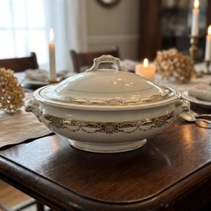 Może przedstawiać: Biała, zakryta miska do serwowania ze złotymi i kwiatowymi akcentami. Naczynie ma uchwyt na pokrywie i dwa uchwyty boczne. Stoi na ciemnym drewnianym stole, z świecami i suszonymi kwiatami w tle.